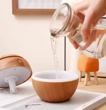 Person pouring liquid from a glass container into a wooden bowl on a light-colored surface.